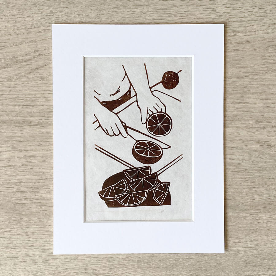 A eco friendly linocut displayed in a conservation mount showing a pair of hands carefully cutting slices of orange fruit. An limited edition linocut by Bert and Roxy