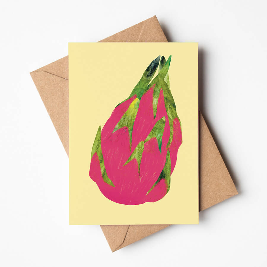 A cheerful eco-friendly greeting card featuring a tropical pink dragon fruit on a yellow background. The card is on top of a brown envelope.