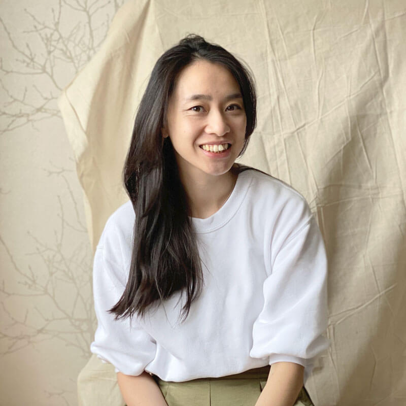 Headshot of printmaker Jessica Yeong of Bert and Roxy, a British Chinese Creative, sitting and smiling against a neutral fabric background 