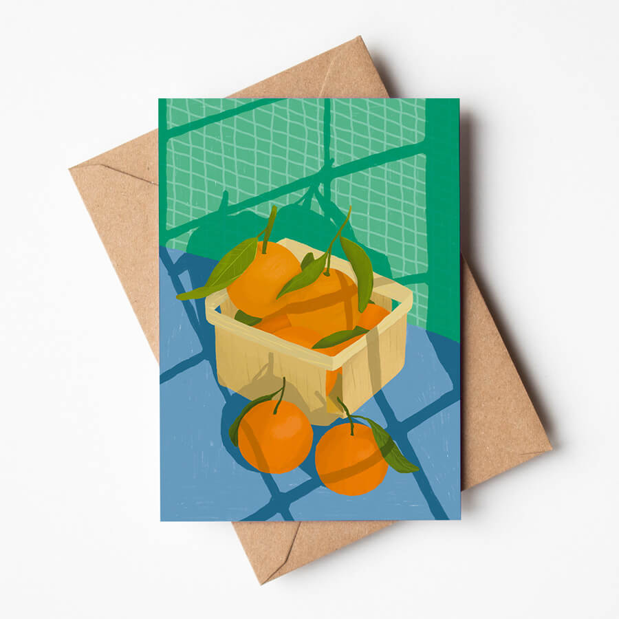 An illustrated greeting card and recycled brown envelope of a punnet of fresh oranges sitting in the sun with overcast shadows from the window