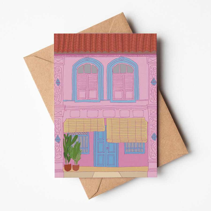 A greeting card, printed on eco friendly materials, displayed on top of a brown envelope. The card has a design of a pink malaysian heritage house