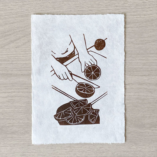 A sustainably made linocut by Jessica Yeong of Bert and Roxy showing a pair of hands cutting orange fruit slices on deckled washi paper