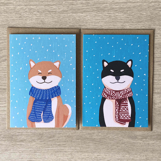 Recycled Christmas Card Pack, featuring A red shiba and a black shiba wrapped up warmly in scarves wishing you Merry Christmas